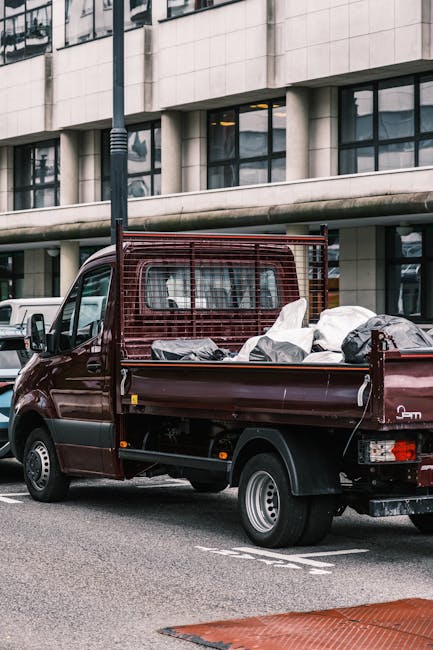 A burgundy flatbed truck parked on an urban street in front of a modern, multi-storey building with large glass windows and a concrete facade. The truck's cargo area is loaded with various rubbish bag