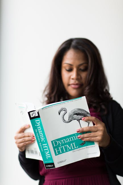 A woman with medium-length dark hair, dressed in a black blazer and a maroon top, is standing indoors against a plain white background. She is holding an open book titled 'Dynamic HTML' with a cover f